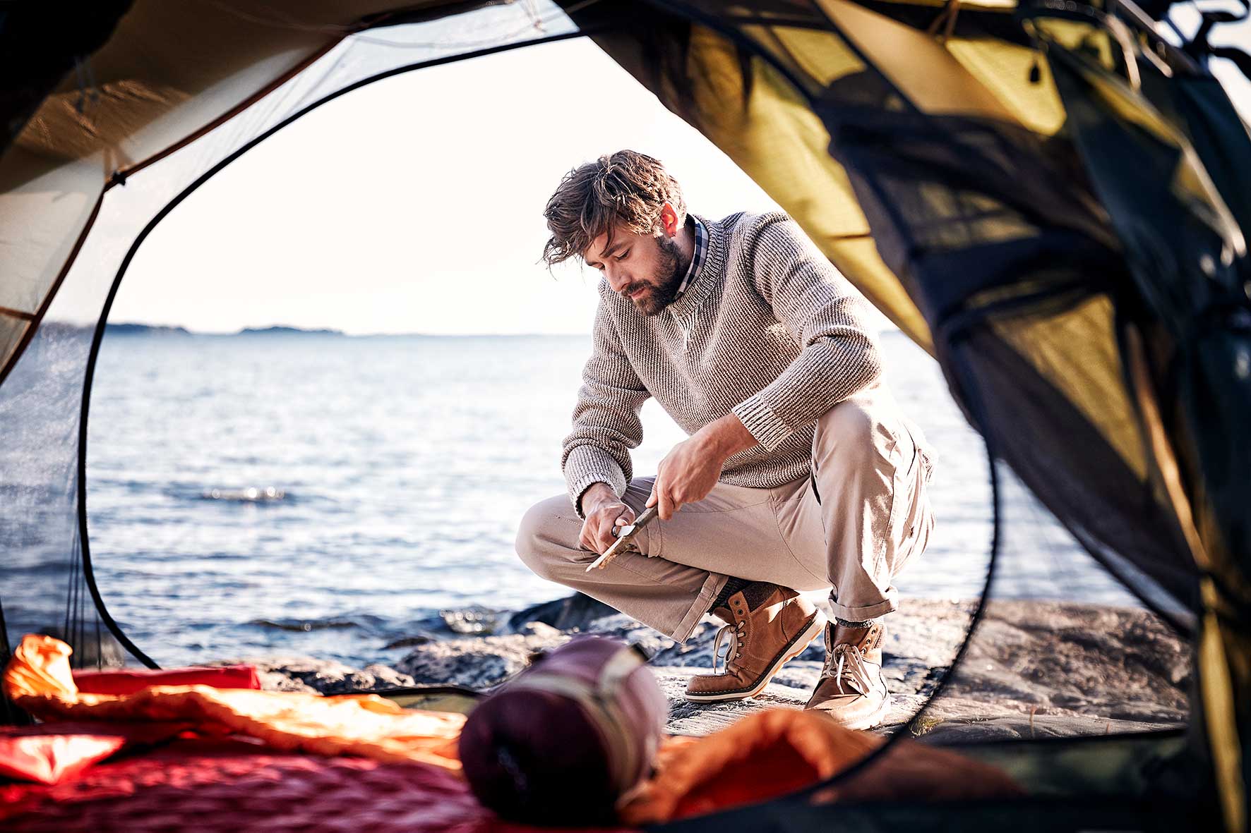 Nicklas crouching on a rock near the sea wearing fair fashion and whittling away at a stick, shot through the inside of his tent.